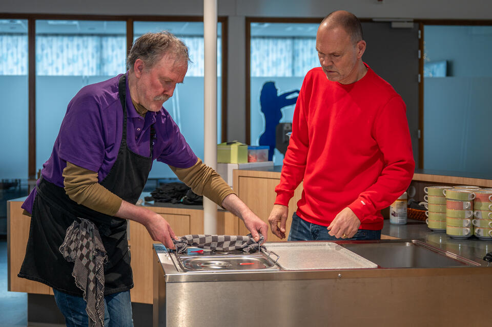 Kantinemedewerker zet een warmhoudschaal in een bak een andere medewerker kijkt ernaar